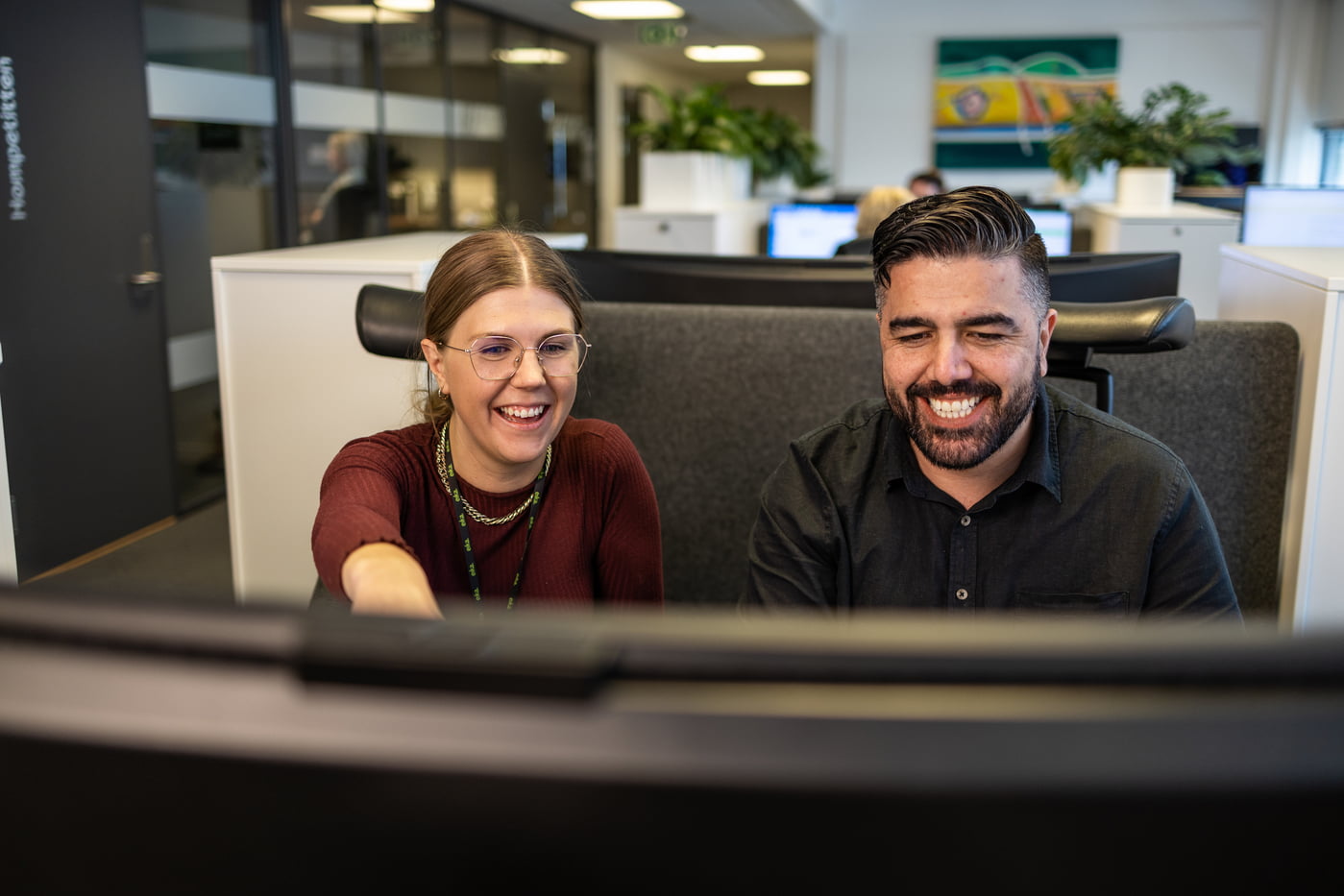 kvinne og mann peker og smiler til pc-skjermen i et åpent kontor landskap